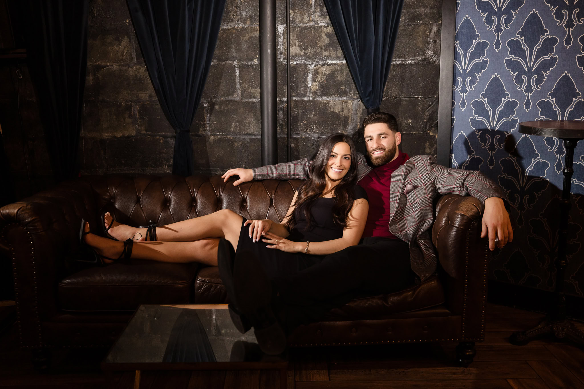 Couple relaxes together on a vintage leather couch at Charlotte’s Speakeasy, surrounded by moody textures and romantic lighting during their Long Island engagement session.