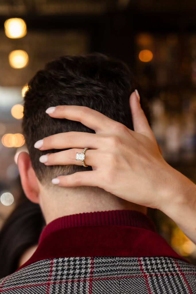 Close-up of bride-to-be’s hand with a radiant ring resting on her fiancé’s neck during their romantic session in Long Island.
