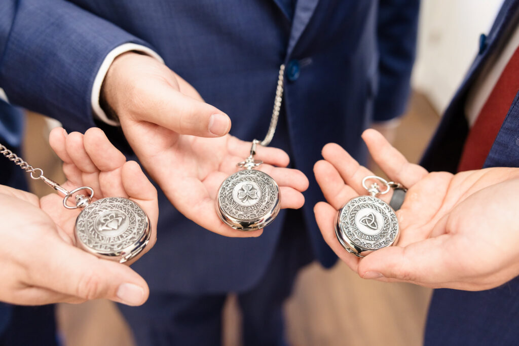 Close-up of groomsmen in blue suits holding engraved silver pocket watches featuring Irish symbols before a wedding.