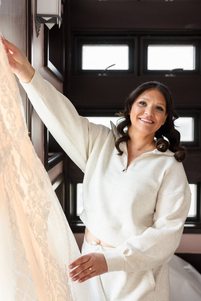 Bride smiling while holding her lace wedding gown during a relaxed morning at home before her fall wedding at Gatsby on the Ocean.