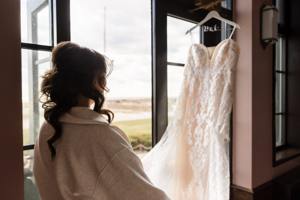 Bride gazes out a window at her lace wedding dress hanging near the waterfront on the morning of her fall wedding at Gatsby on the Ocean.