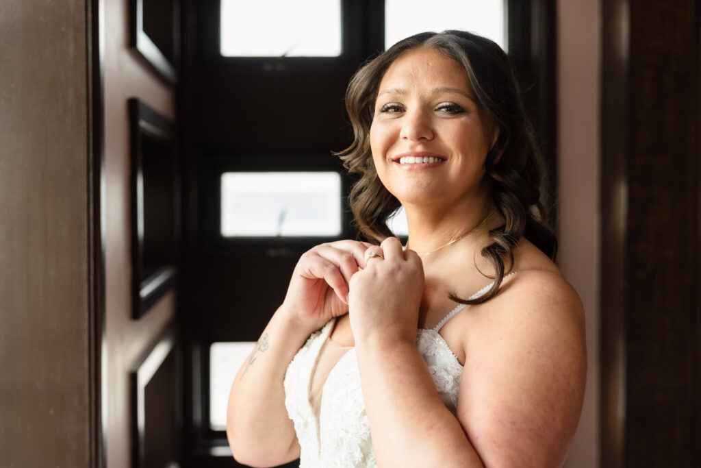 Bride smiles while adjusting her necklace in natural light, wearing a lace gown on the morning of her fall wedding at Gatsby on the Ocean.