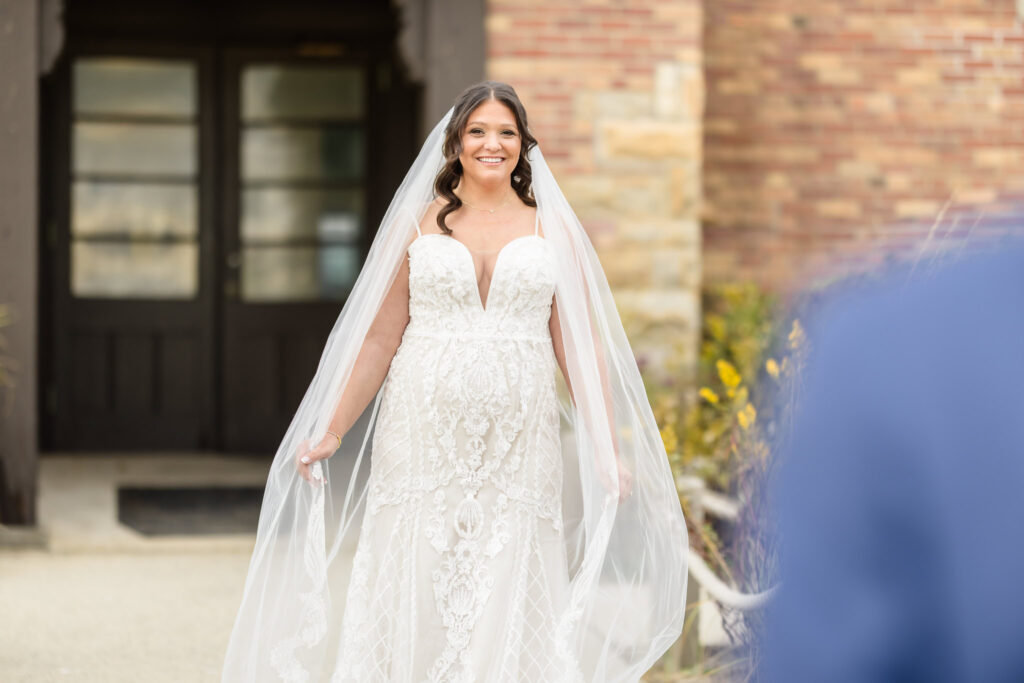 Bride smiles radiantly in her lace gown and flowing veil during a joyful first look outside Gatsby on the Ocean.