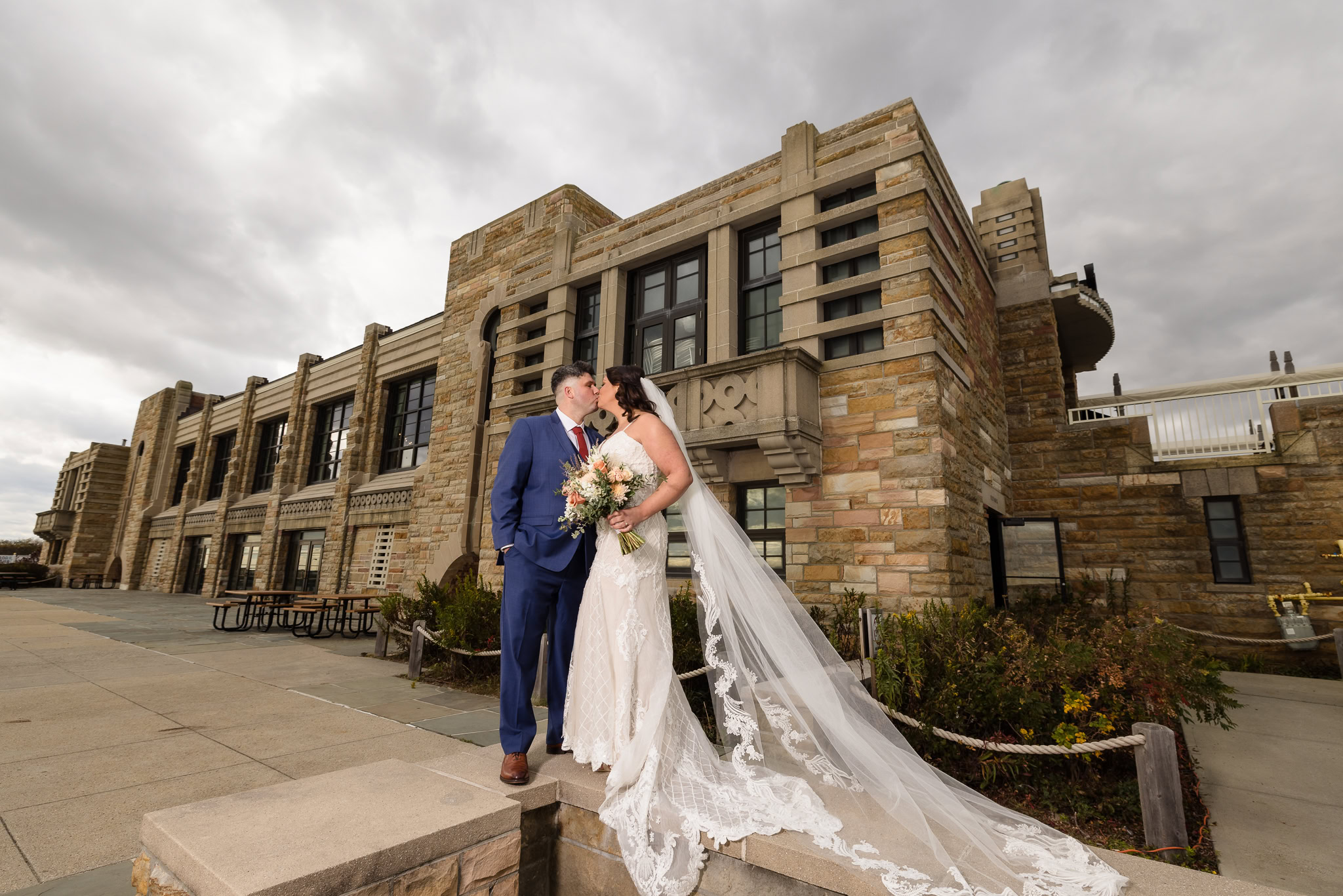 Bride and groom share a romantic kiss in front of the historic Gatsby on the Ocean building at Jones Beach, her lace-trimmed veil flowing dramatically in the wind beneath a moody sky.