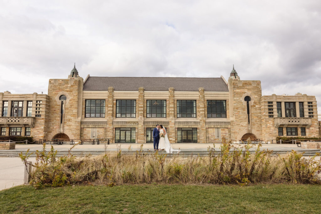Bride and groom stand in a sweet embrace in front of the grand, historic Gatsby on the Ocean venue at Jones Beach, with its symmetrical stone facade and dramatic architecture under a cloudy sky.