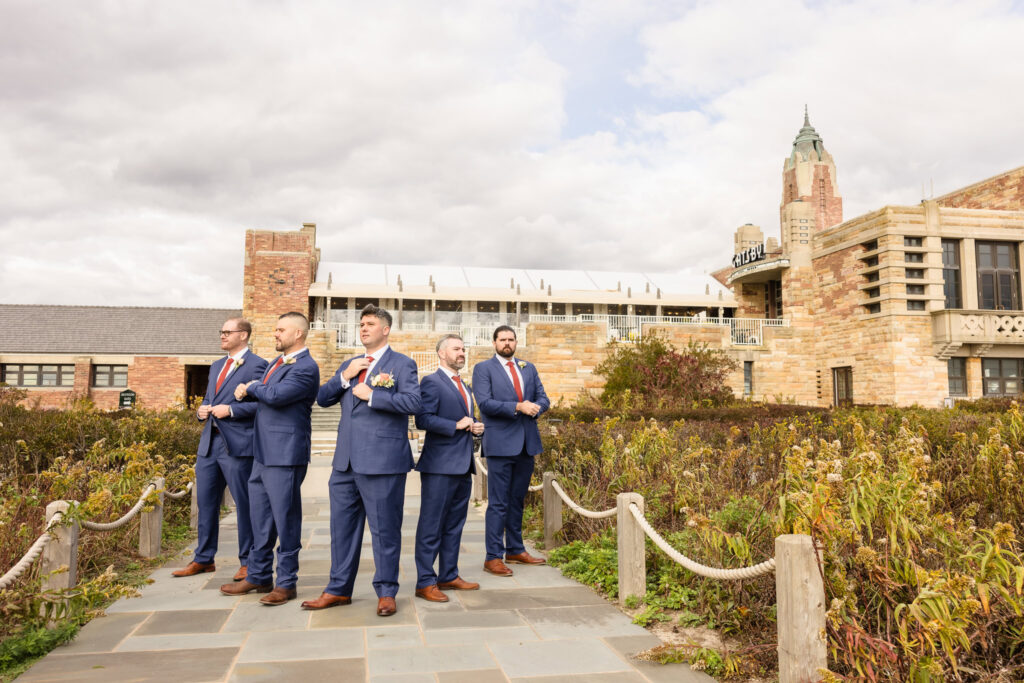 Groom and groomsmen pose confidently in matching blue suits with red ties in front of the iconic Gatsby on the Ocean venue at Jones Beach, exuding style and camaraderie under a partly cloudy sky.