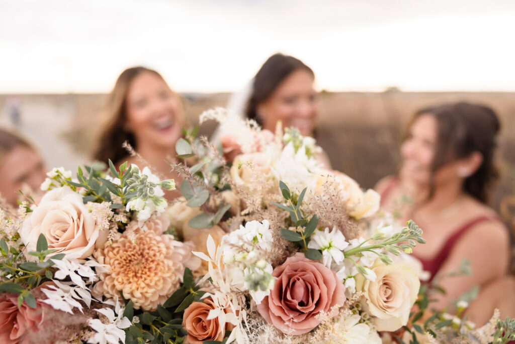 A vibrant close-up of wedding bouquets featuring blush, ivory, and peach-toned flowers with greenery, with the smiling, out-of-focus bridesmaids and bride joyfully gathered in the background.