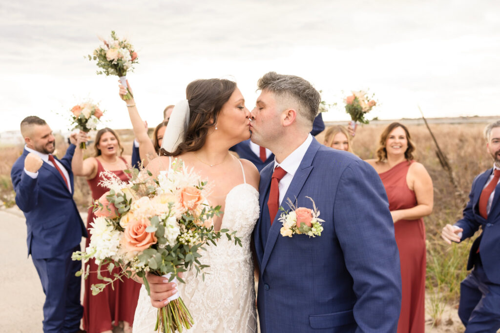 The bride and groom share a kiss while holding a floral bouquet, surrounded by a cheering bridal party dressed in blue suits and rust-colored dresses, celebrating joyfully in an outdoor setting with a natural backdrop.