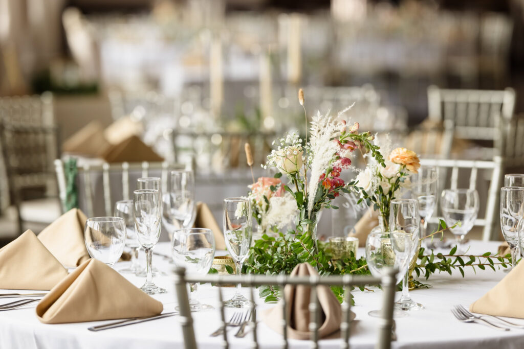 A close-up of an elegantly set wedding reception table featuring champagne flutes, water glasses, beige folded napkins, and a lush floral centerpiece with greenery and soft-colored blooms, surrounded by white chairs.