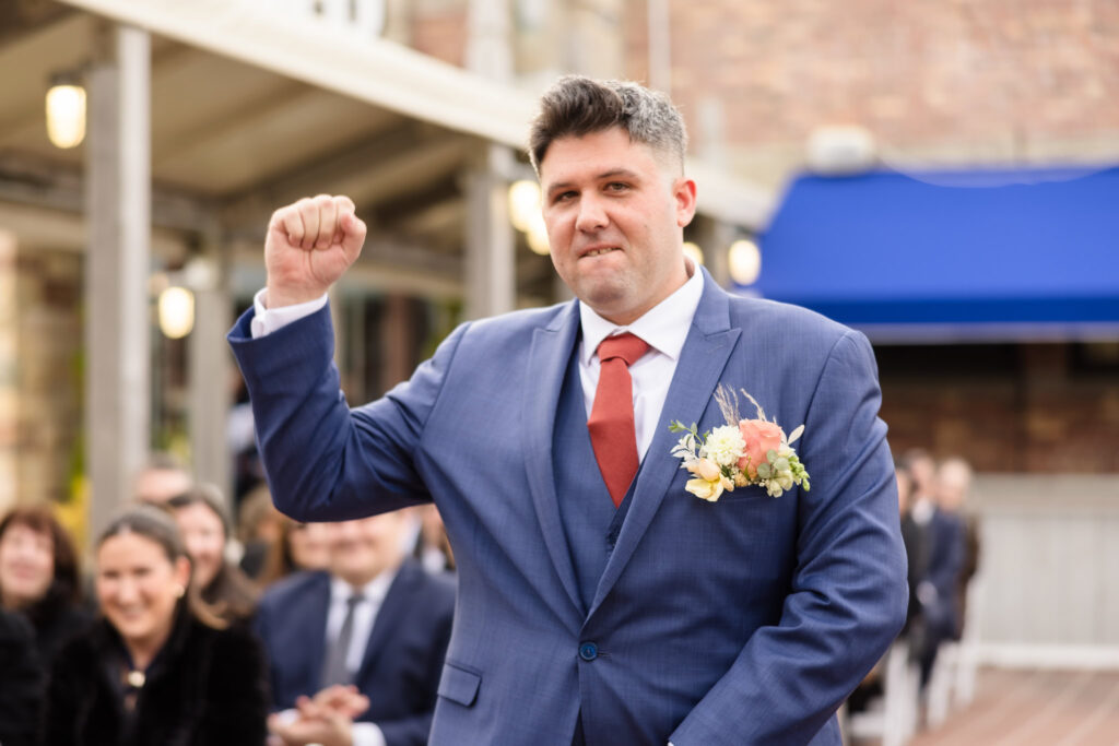 A groom in a blue suit with a red tie and floral boutonniere raises his fist in excitement as he walks down the aisle, smiling with guests seated and watching in the background.