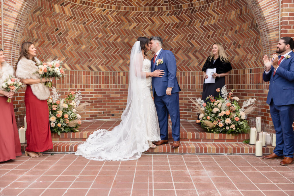 Bride and groom share their first kiss during a fall wedding ceremony on the East Patio at Gatsby on the Ocean, surrounded by peach florals and brick architecture.