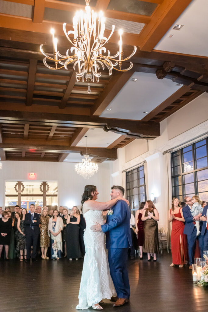 Bride and groom share their first dance under chandeliers at Gatsby on the Ocean, surrounded by loved ones during their intimate fall wedding reception.