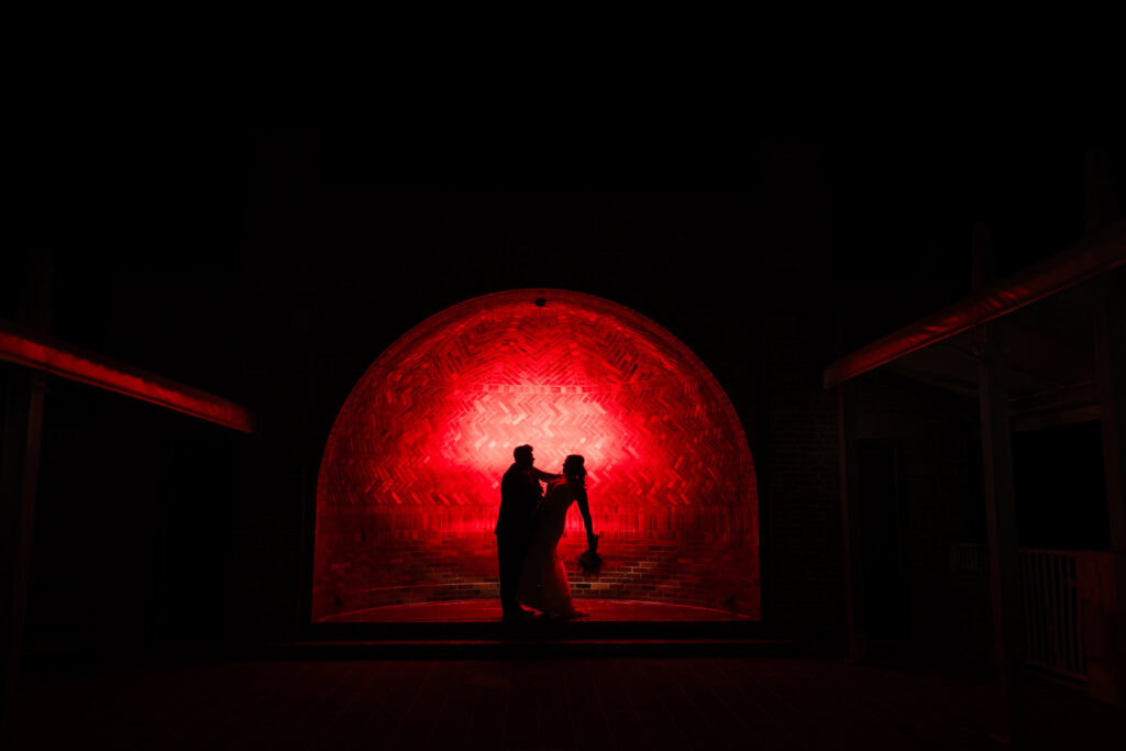 Dramatic nighttime silhouette of bride and groom in front of red-lit brick archway at Gatsby on the Ocean during their fall wedding.