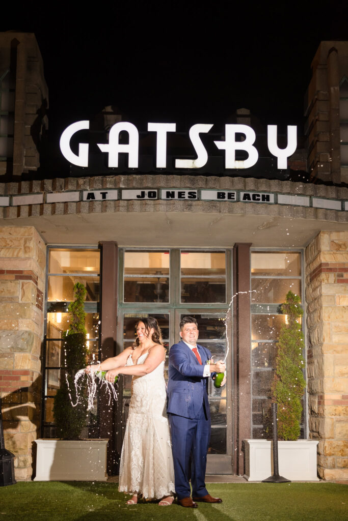 Bride and groom pop champagne in celebration under the glowing Gatsby on the Ocean sign at Jones Beach during their fall wedding night.