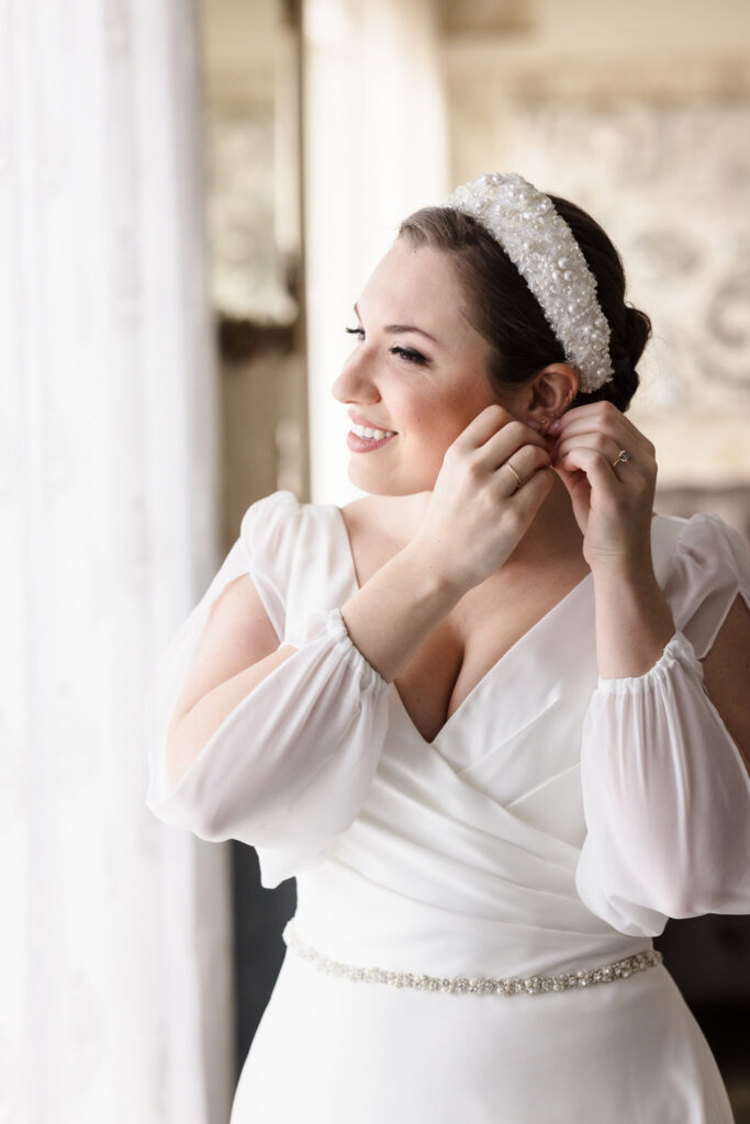 Bride getting ready, smiling as she puts on earrings in natural window light, wearing a pearl headband and flowy-sleeved gown.