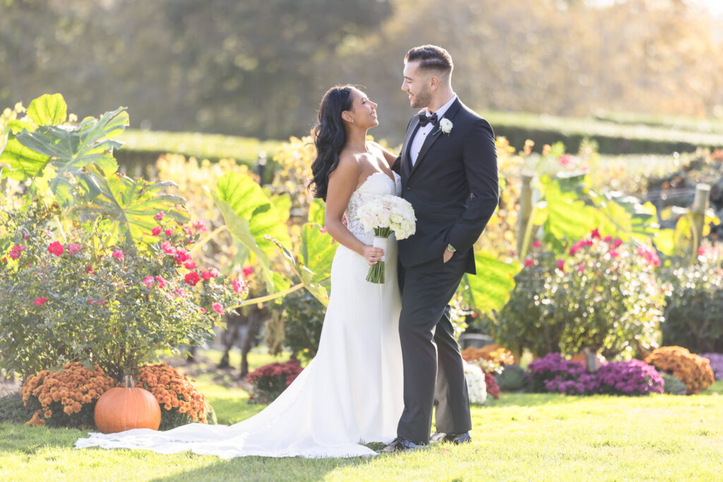 Bride and groom smiling at each other in Giorgio’s Baiting Hollow garden, surrounded by vibrant fall flowers, pumpkins, and lush greenery.