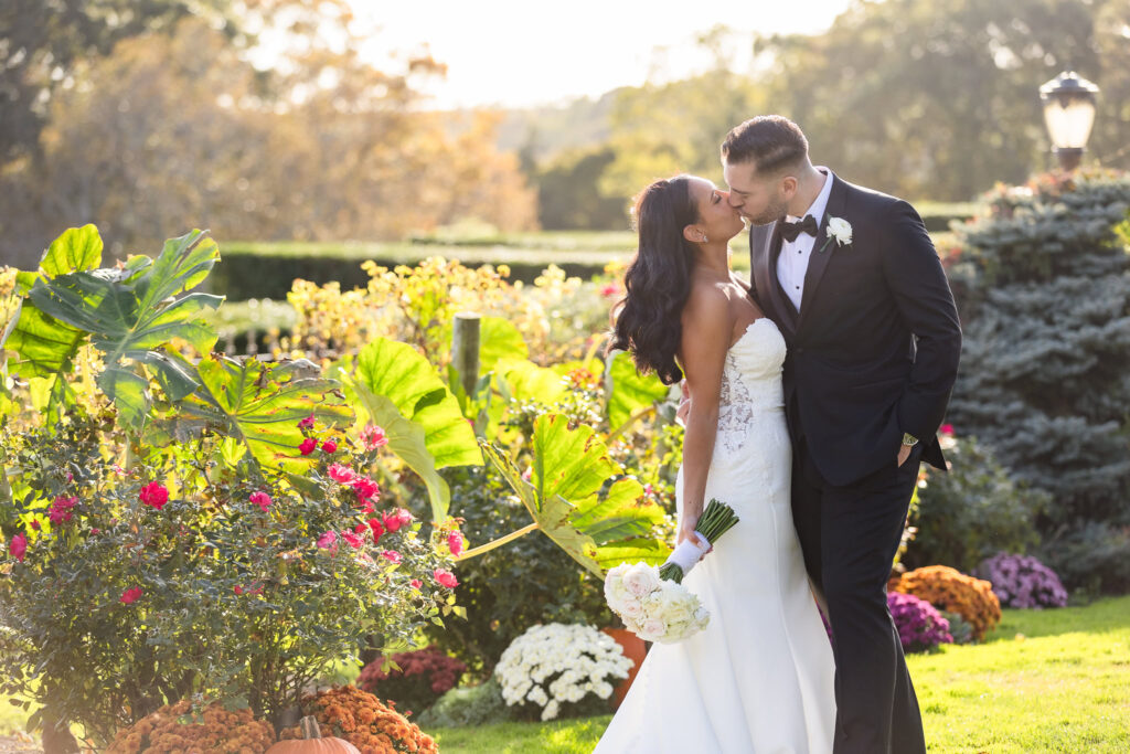Bride and groom sharing a kiss in the vibrant gardens at Giorgio’s Baiting Hollow, surrounded by fall florals, greenery, and pumpkins.