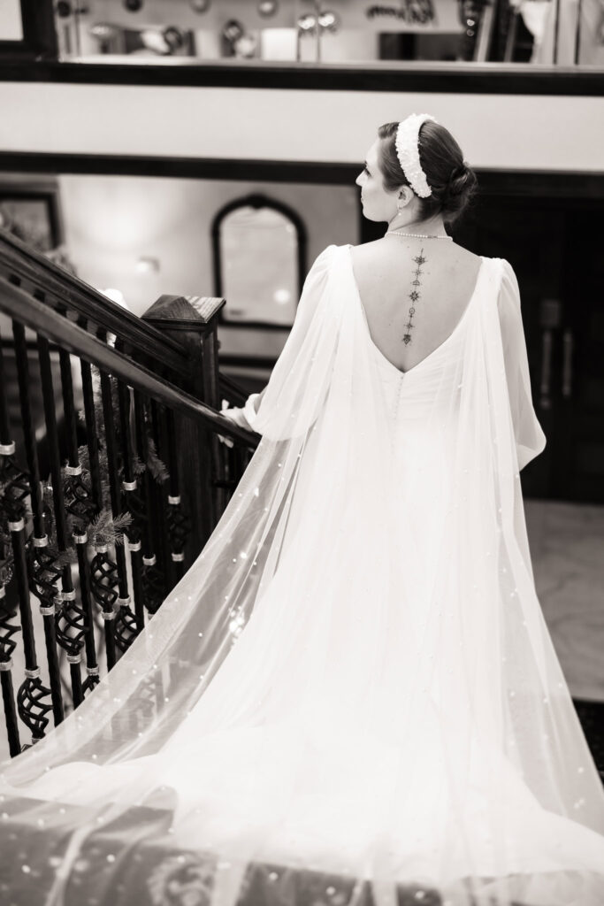 Black and white portrait of a bride descending the staircase at Giorgio’s Baiting Hollow, showcasing her beaded cape veil and back tattoo.