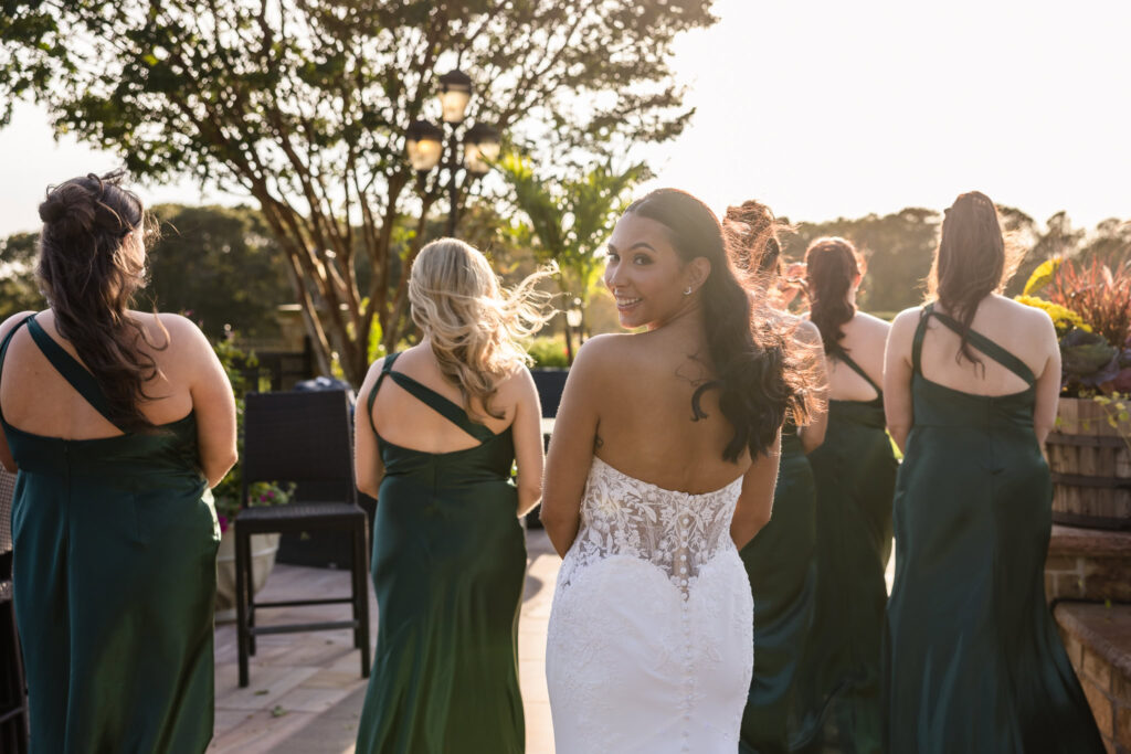 Bride smiling over her shoulder as she walks with bridesmaids in emerald green dresses at Giorgio’s Baiting Hollow during golden hour.