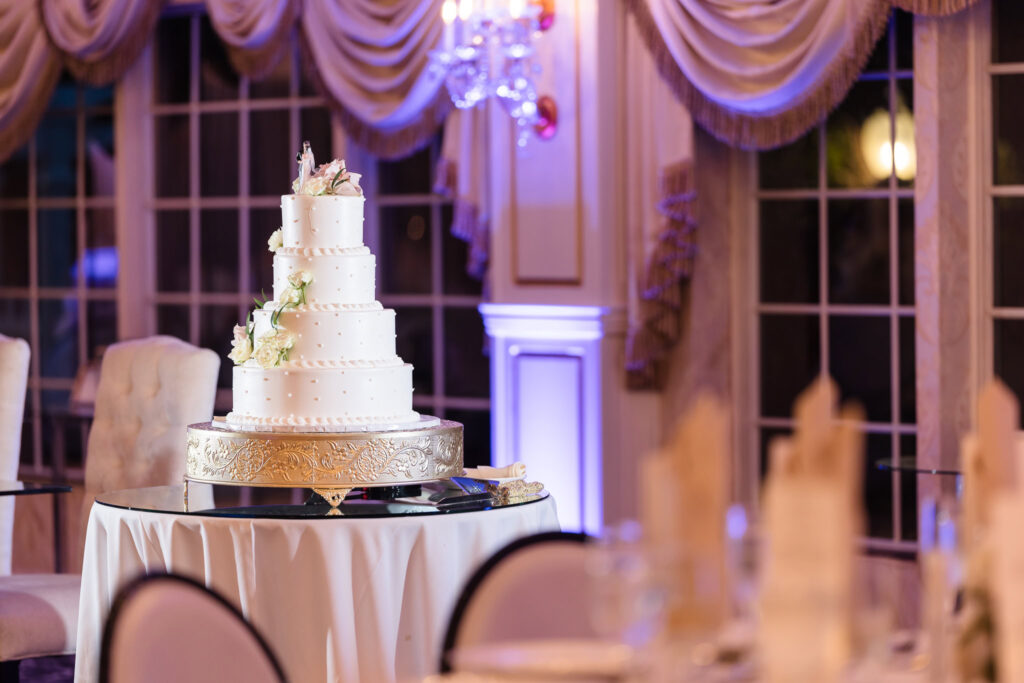 Elegant five-tier white wedding cake with floral accents on a gold stand, displayed in the ballroom at Giorgio’s Baiting Hollow.