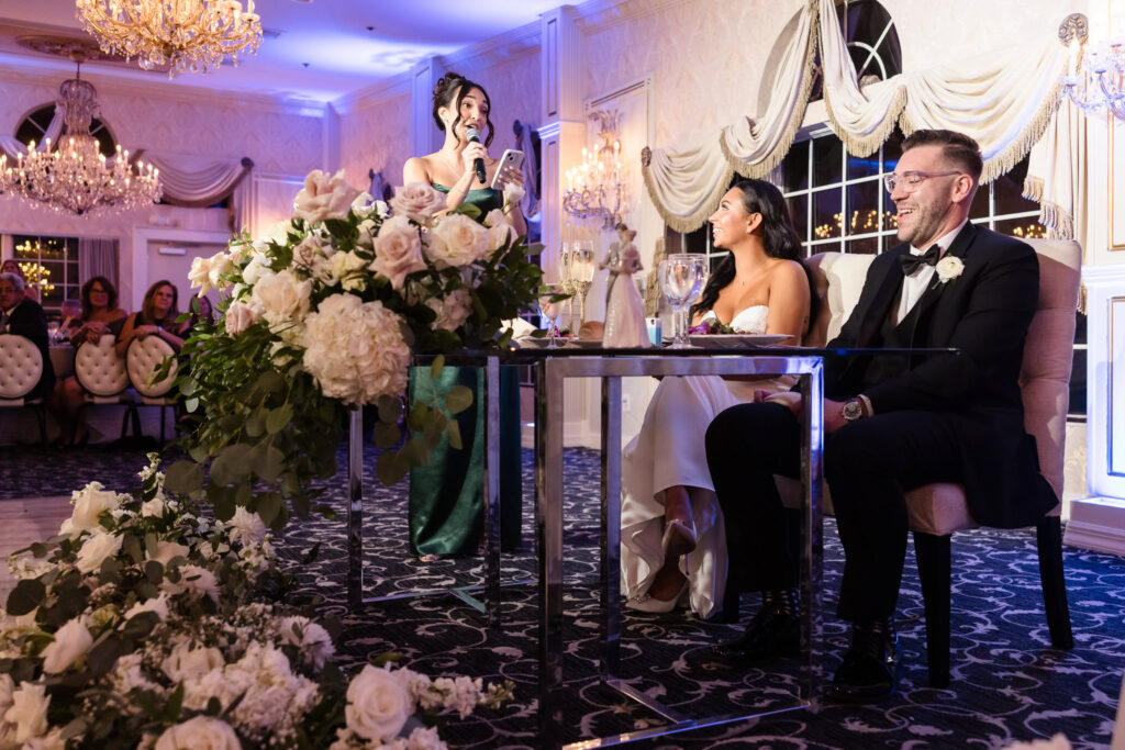 Maid of honor giving a heartfelt speech as the bride and groom laugh at the sweetheart table adorned with white floral arrangements at Giorgio’s Baiting Hollow.