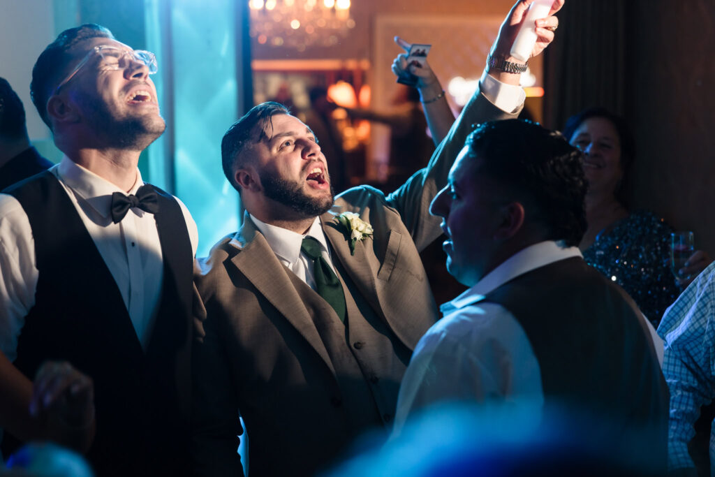 Groom and guests singing and celebrating on the dance floor under colorful lighting during the wedding reception.