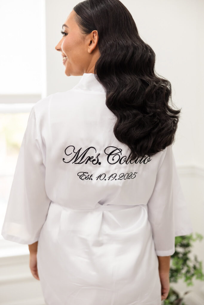 Bride AnnaLisa wearing a white robe embroidered with “Mrs. Coletto, Est. 10.19.2025” while getting ready for her fall wedding, showcasing elegant bridal beauty and personal details.