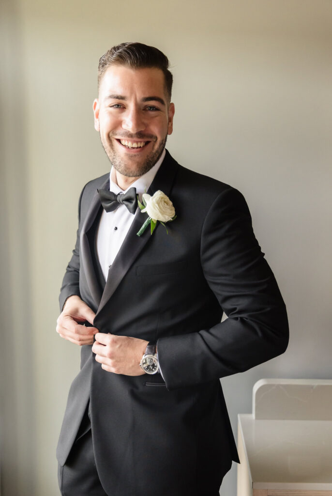 Groom Marc smiling as he buttons his classic black tuxedo, wearing a white rose boutonniere and preparing for his fall wedding with timeless elegance.