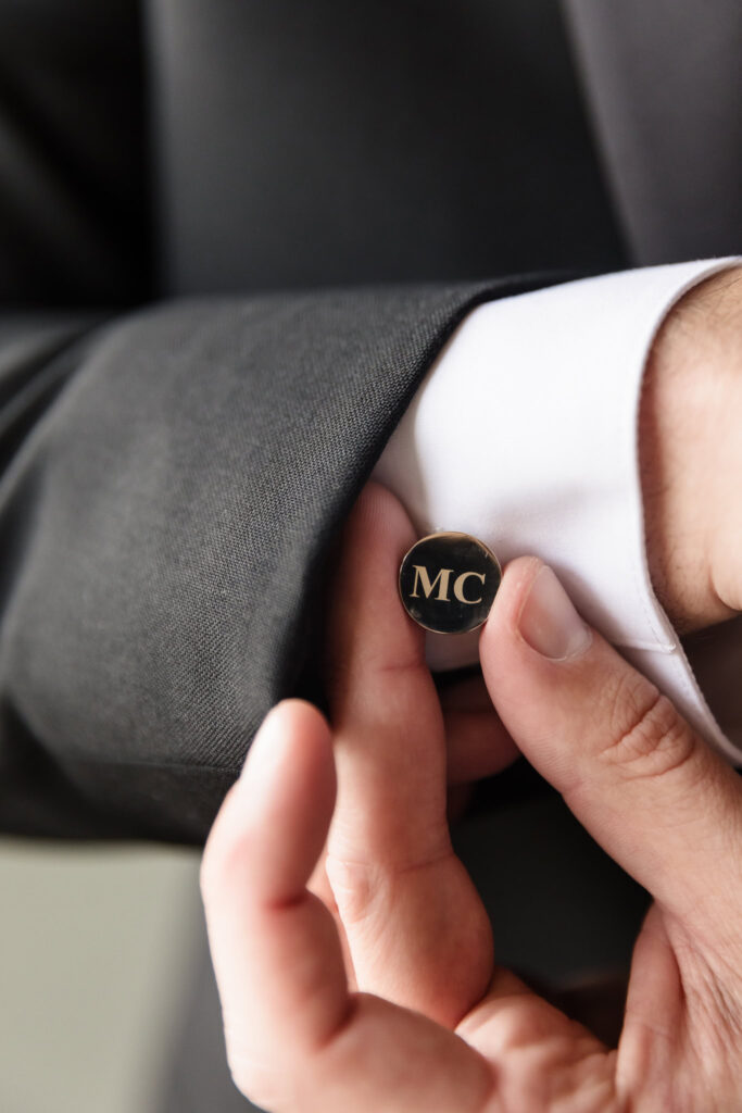 Close-up of groom Marc fastening a personalized black and gold cufflink engraved with his initials “MC,” adding a refined touch to his wedding attire.