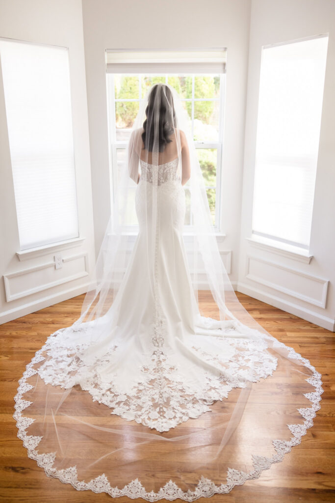 Back view of bride AnnaLisa standing in front of a bright window, showcasing the intricate lace train of her veil and gown on the morning of her elegant wedding.