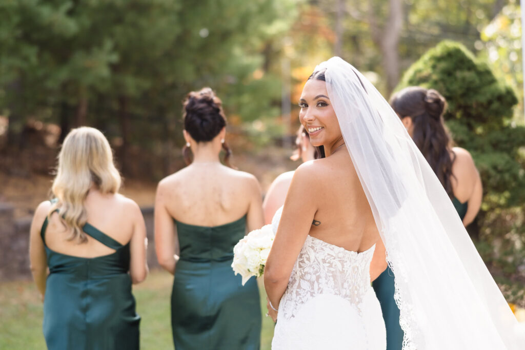 Bride AnnaLisa smiles over her shoulder while walking with her bridesmaids in emerald green gowns, her lace wedding dress and veil flowing in the autumn light at Giorgio’s Baiting Hollow.