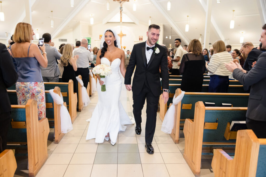 Newlyweds AnnaLisa and Marc walk joyfully down the aisle at St. Mark’s Church as guests cheer and capture the moment, just before heading to their Giorgio’s Baiting Hollow wedding celebration.
