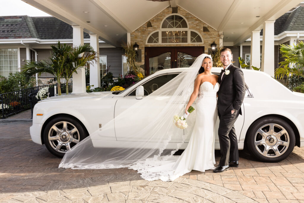 Bride and groom pose in front of a classic white Rolls Royce at Giorgio’s Baiting Hollow, with AnnaLisa’s cathedral veil flowing beside her and the venue’s elegant entrance in the background.