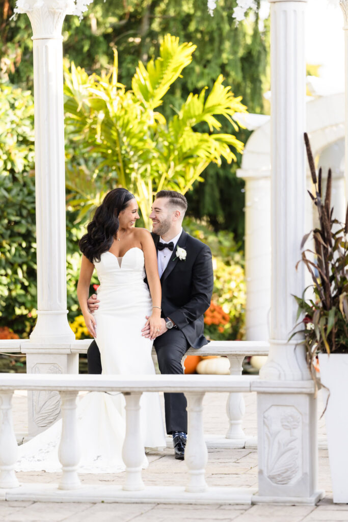Bride and groom sit together under the white stone gazebo at Giorgio’s Baiting Hollow, sharing a joyful moment surrounded by greenery and fall florals during their elegant autumn wedding.