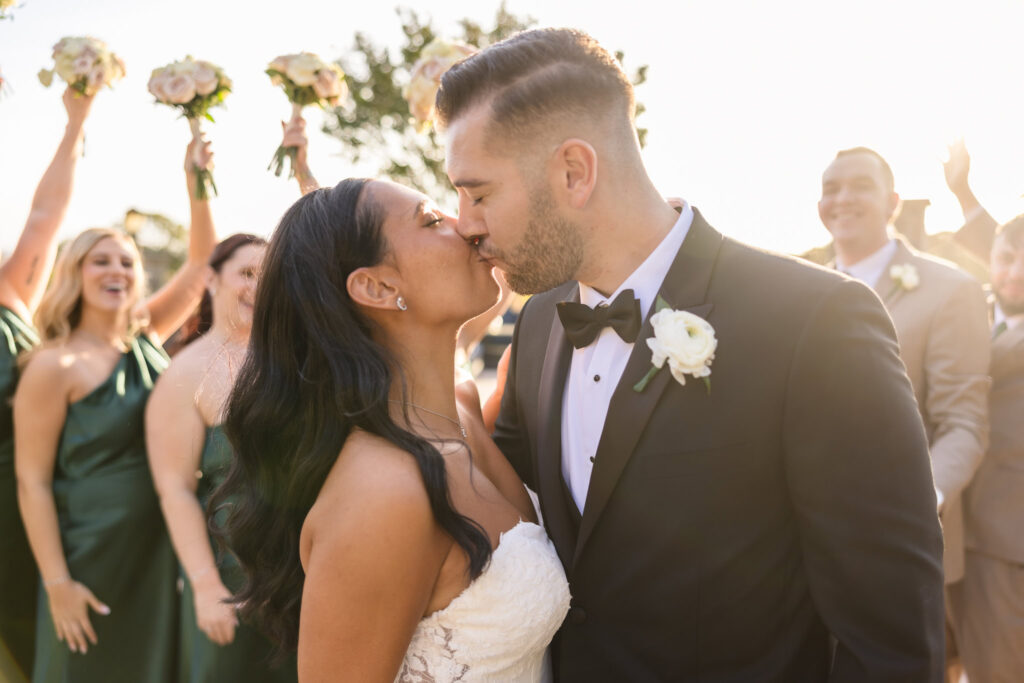 Bride and groom share a celebratory kiss surrounded by their cheering wedding party at Giorgio’s Baiting Hollow, with golden sunlight and joyful energy filling the frame.