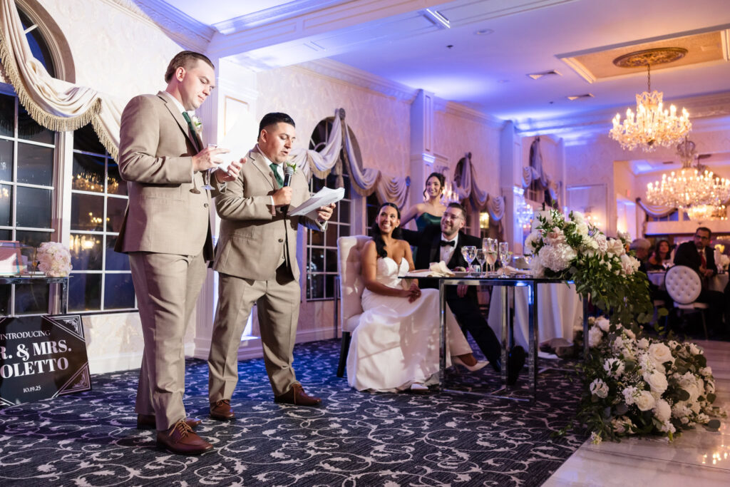 The best men deliver a heartfelt and humorous toast as AnnaLisa and Marc smile at their sweetheart table, surrounded by floral arrangements and warm candlelight at their Giorgio’s Baiting Hollow reception.