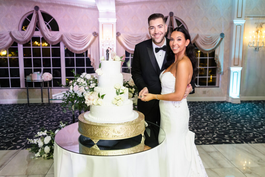 Bride and groom pose with smiles as they prepare to cut their elegant white wedding cake at Giorgio’s Baiting Hollow, surrounded by floral arrangements and reception decor.