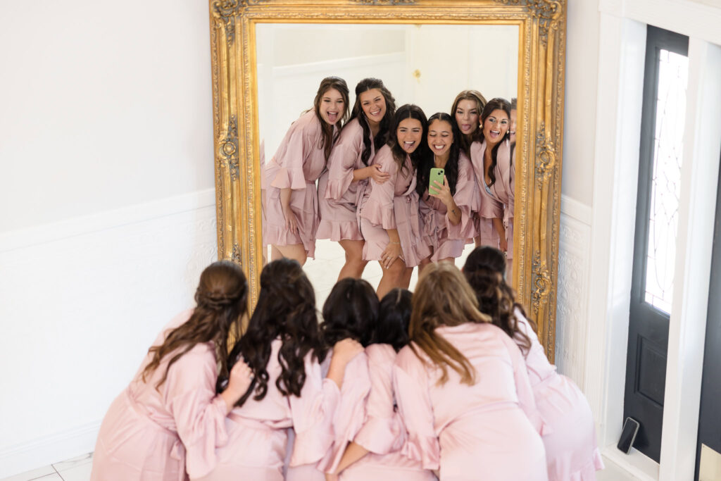Bride and bridesmaids in matching blush robes pose playfully in front of a gold-framed mirror at the bridal suite inside Hiland Park Wedding Venue in Queensbury, NY.