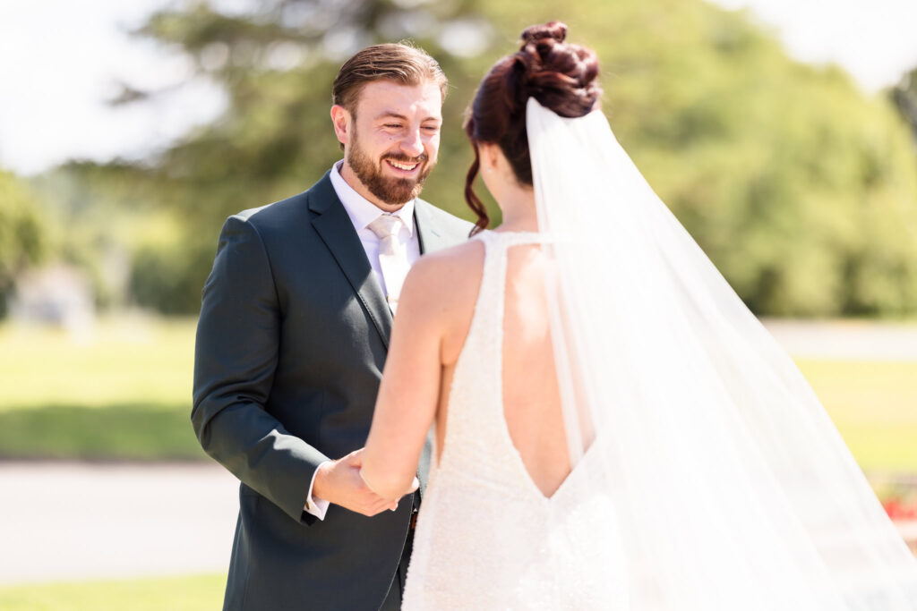 Groom smiles warmly at his bride during their first look outside at Hiland Park Wedding Venue, surrounded by greenery and soft natural light in Queensbury, NY.