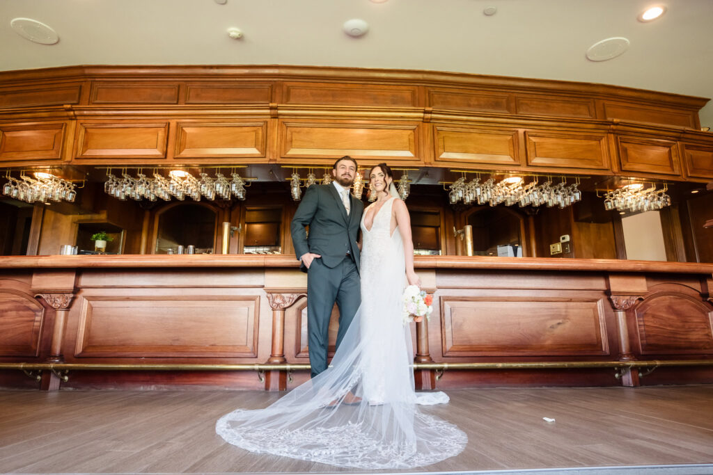 Bride and groom pose together in front of the grand wooden bar at Hiland Park Country Club, with the bride’s long veil elegantly trailing across the floor.