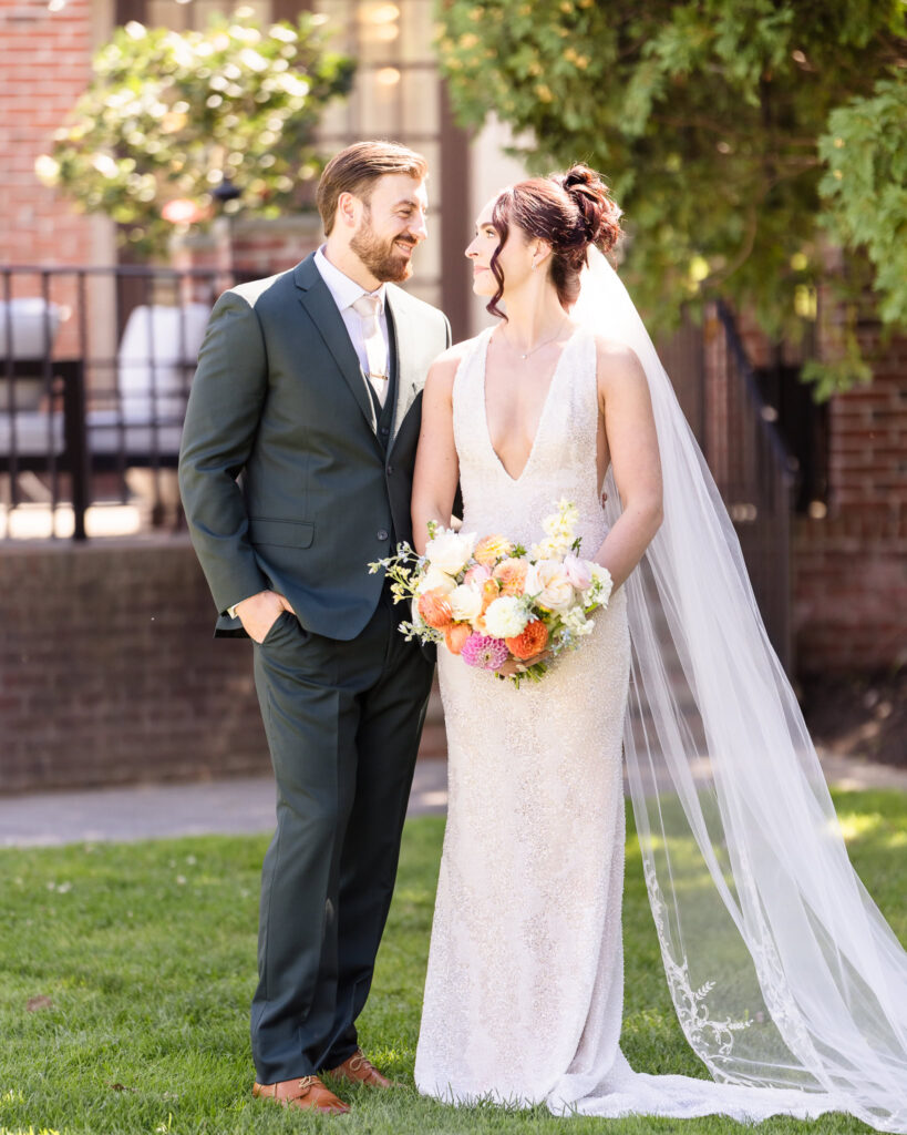 Bride and groom gaze at each other lovingly in the garden at Hiland Park Wedding Venue, with lush greenery and brick architecture in the background.