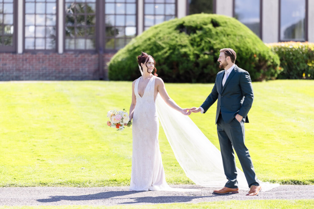 Bride and groom hold hands and smile at each other while walking the lush grounds of Hiland Park Wedding Venue, with elegant windows and trimmed greenery behind them.