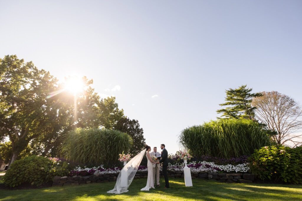 Bride and groom exchange vows during an outdoor ceremony at Hiland Park Wedding Venue, surrounded by lush greenery, vibrant florals, and golden sunlight.