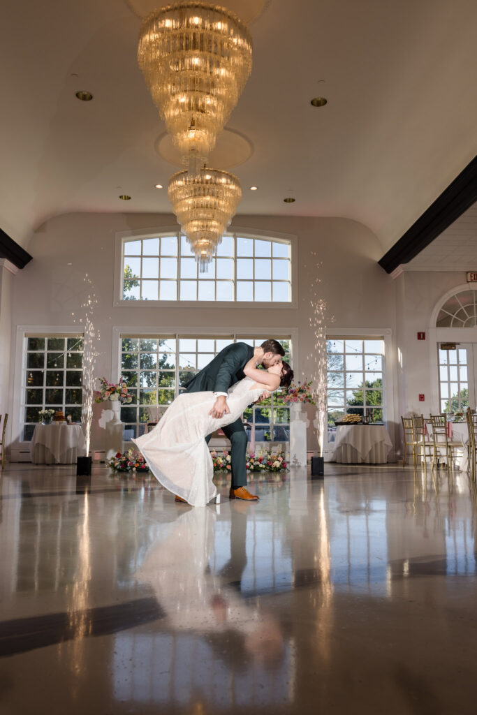 Groom dips and kisses bride on the dance floor during their first dance at Hiland Park’s grand ballroom, with chandeliers and sparkler fountains enhancing the moment.