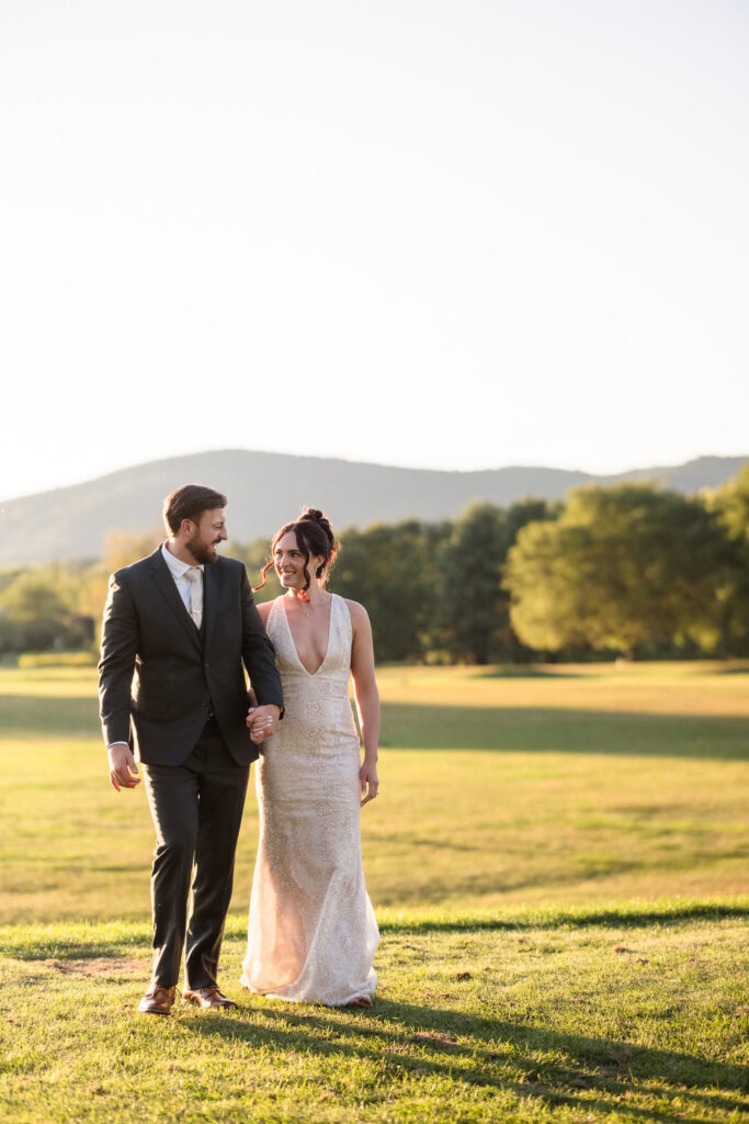 Bride and groom smile at each other while walking hand-in-hand through the sunlit fields of Hiland Park Wedding Venue, with scenic mountains in the background.