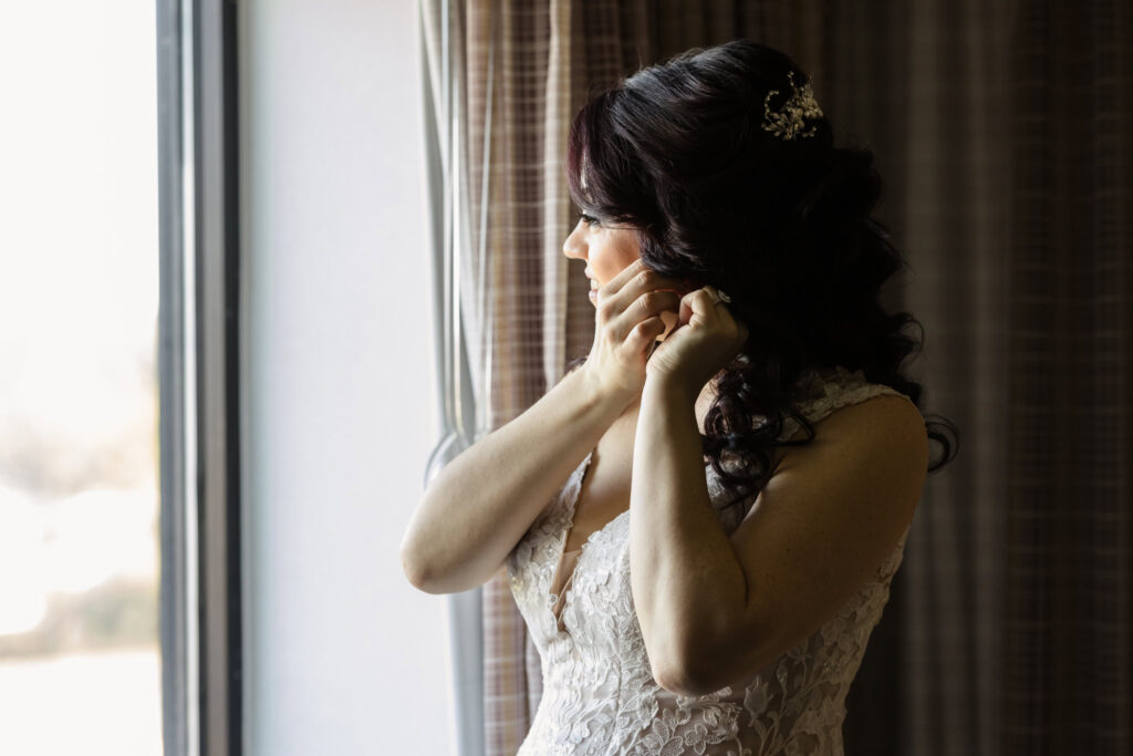 Bride putting on earrings by the window in soft morning light, wearing a lace wedding gown with floral appliqué and elegant curled hair.
