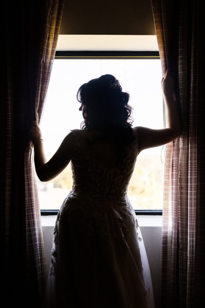 Silhouette of bride in lace gown standing at the window, holding open the curtains as soft daylight frames her during a quiet pre-ceremony moment.