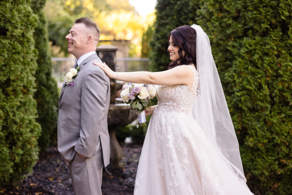 Bride in lace gown reaching to tap groom’s shoulder during their first look in the gardens at Larkfield, capturing a joyful and emotional pre-ceremony moment.