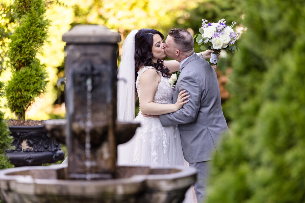 Bride and groom share a kiss by the garden fountain at Larkfield, with the bride lifting her bouquet of white and lavender roses in celebration.