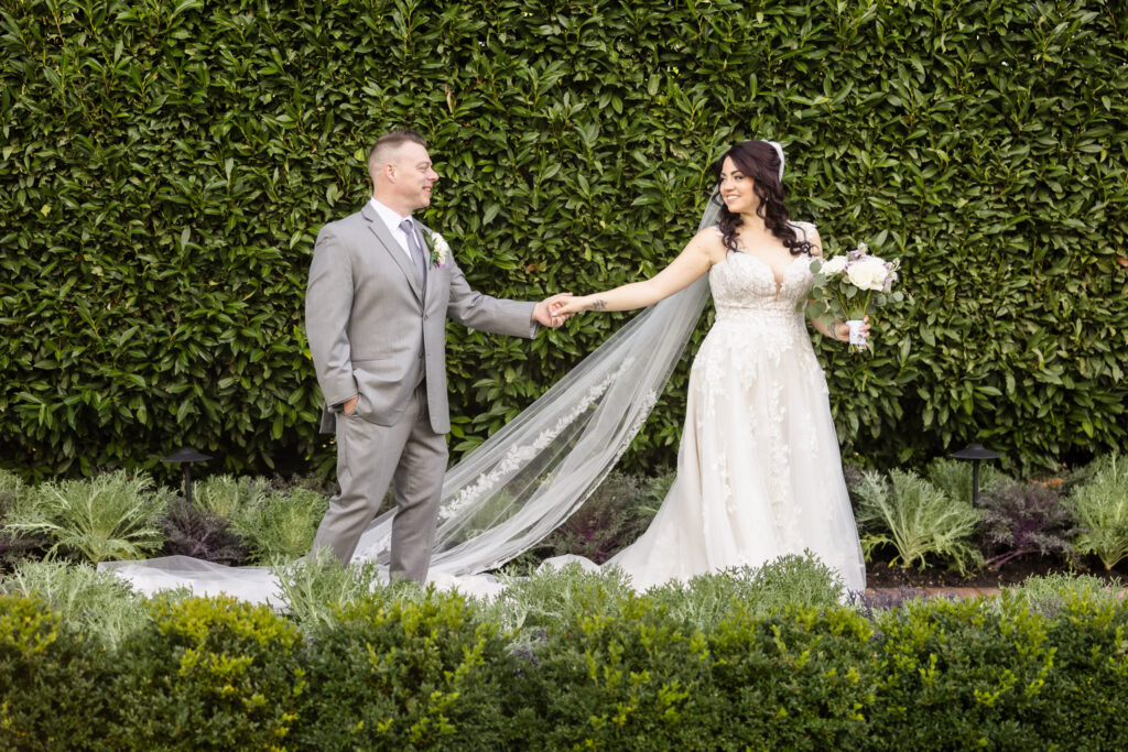 Bride and groom holding hands and smiling in front of lush greenery at Larkfield, with her long lace veil trailing behind and bouquet in hand.